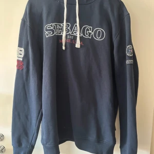 Svart hoodie från Sebago - Säljer en svart hoodie från Sebago med tryck på bröstet och ärmen. Den har en justerbar huva med snören och ribbade muddar. Perfekt för en avslappnad stil.