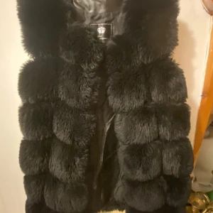 Svart pälsväst - Snygg svart pälsväst med fluffig textur och elegant design. Perfekt för att hålla sig varm och stilren under kyliga dagar. Västen har en lyxig känsla och passar bra till olika outfits.
