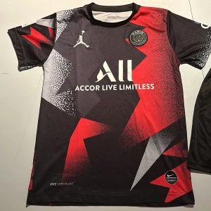 Svart och röd PSG fotbollströja med shorts - Snygg PSG fotbollströja med Mbappé 7 på ryggen. Tröjan har ett coolt mönster i svart, rött och vitt med korta ärmar. Tillhörande svarta shorts med PSG-logga. Ganska liten i storleken. Perfekt för fotbollsfans!