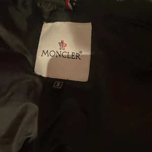 Svart vest med glans från moncler  - Snygg svart vest från Moncler med klassisk design. Vest har en unik etikett med serietidningsmotiv på insidan. Perfekt för halv kyliga dagar med stil.