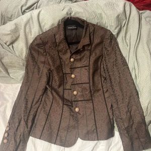 Brun kavaj 70-tal - Elegant brun kavaj från Marcona med ett subtilt mönster och dekorativa knappar framtill. Kavajen har en klassisk krage och långa ärmar, perfekt för en stilren look.