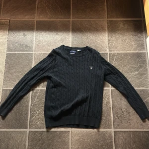 Svart kabelstickad tröja från GANT - Säljer en stilren svart kabelstickad tröja från GANT i premium bomull. Tröjan har en klassisk design med rund hals. Knappt använd och den passar M. 