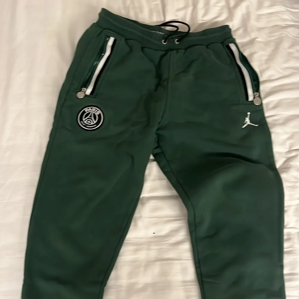 Snygga gröna mjukisbyxor med Paris Saint-Germain-logga och Jumpman-märke. Byxorna har dragkedjor vid fickorna och snörning i midjan för justerbar passform. Perfekta för en avslappnad stil.. Farkut & Housut.