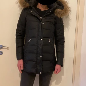 Svart parkas  med pälsdetaljer - Snygg svart dunjacka med huva och pälsdetaljer. Jackan har dragkedja och knappar framtill samt praktiska fickor med dragkedja. Perfekt för kalla dagar med sin långa passform och vadderade design.