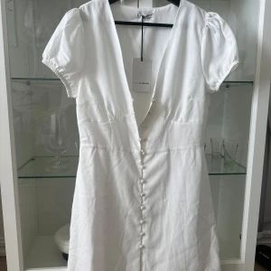 Vit klänning från GlamorousXnelly - Elegant vit klänning från GlamorousXnelly med korta ärmar och v-ringning. Klänningen har knappar framtill som ger en stilren look. Perfekt för somriga tillfällen.😍