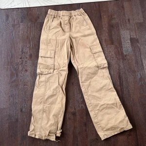 Beige cargopants med fickor - Säljer ett par beige cargopants med flera praktiska fickor och elastisk midja. Byxorna har en avslappnad passform och är perfekta för en casual look. Det finns lite tecken på användning som en liten fläck, som annars inte syns när man har på dem, och lite slitage vid foten. Hör gärna av er vid frågor och funderingar 💕💕