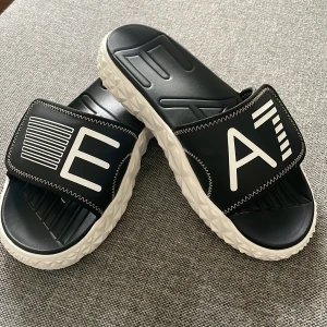 Svarta EA7 sandaler med vit sula - Snygga svarta sandaler med en vit, mönstrad sula. De har en bred rem över foten med vita grafiska detaljer och synliga sömmar. Perfekta för en avslappnad stil.
