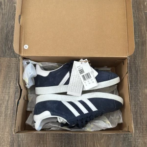 Adidas Gazelle i mörkblå mocka -  Adidas Gazelle sneakers i mörkblå mocka, storlek 38 2/3, med vita detaljer och de klassiska tre ränderna på sidan. Skorna har snörning och en vit sula. Aldrig använda utomhus, endast testat dom en gång. Säljer dessa eftersom de var för små för mig. Nypris 1200 kr, säljer nu för 650.