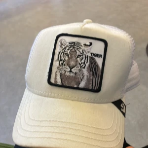 Vit keps med tigerbroderi - Snygg vit keps med ett coolt broderi av en tiger på framsidan. Kepsen har ett nätmaterial på baksidan för bättre ventilation och en justerbar rem för perfekt passform. Perfekt för en avslappnad stil.