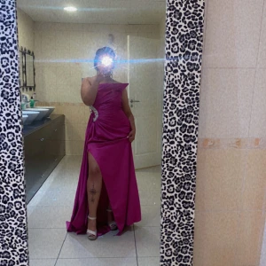 Rosa axelbandslös långklänning - Snygg rosa långklänning med en axelbandslös design och en elegant slits. Klänningen har en glittrig detalj vid midjan som ger extra glamour. Perfekt för en festlig kväll!