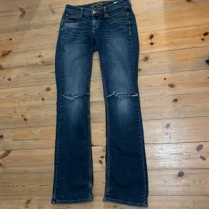 Blå bootcut jeans från Arizona - Snygga blå bootcut jeans från Arizona med slitna detaljer på knäna. Perfekta för en avslappnad stil. Klassisk femficksdesign och knappgylf. Passar bra till både sneakers och stövlar.