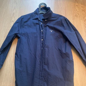 Mörkblå skjorta från GANT - Säljer en stilren mörkblå skjorta från GANT. Skjortan har långa ärmar och knappar framtill. Perfekt för en klassisk look. Storlek 176 16år men är ganska stor i storlek