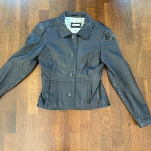Svart skinnjacka från Vila - Snygg svart skinnjacka från Vila i storlek L. Jackan har en klassisk krage och knäppning framtill med knappar. Perfekt för en stilren look med långärmade ärmar och en figurnära passform. Finns tecken på användning men fortfarande bra skick 