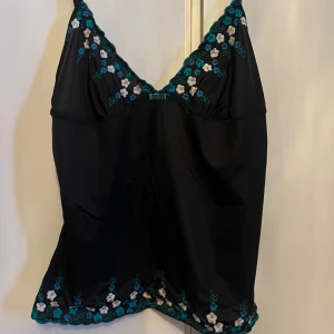 Svart linne med blommig brodyr - Snyggt svart linne med tunna axelband och v-ringning. Detaljerad med vacker blommig brodyr i blått och vitt längs halsringningen och nederkanten. Perfekt för en somrig look! Lappen är bortklippt och är köpt second hand, men skulle gissa på stolek S/M!🌸