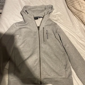 Grå zip hoodie från Sail Racing - Säljer en grå zip hoodie från Sail Racing med dragkedja och huva. Hoodien har en broderad logotyp på bröstet och är tillverkad i ett mjukt material. Perfekt för kyligare dagar.