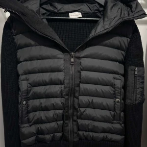 Svart jacka från Moncler - Snygg svart jacka från Moncler med quiltad design och dragkedja. Jackan har en huva och är långärmad med en ficka på ärmen. Perfekt för kyligare dagar.