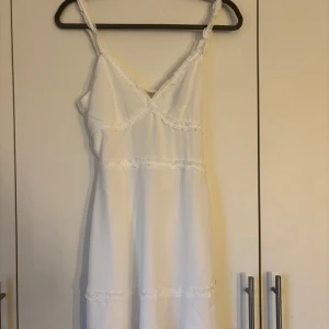 Vit klänning från H&M Divided - Säljer en söt vit klänning från H&M Divided med smala axelband och knytning i ryggen. Klänningen har en v-ringad design och är perfekt för varma dagar. Den är tillverkad i ett lätt och luftigt material. den är öppen i ryggen och använd en gång.