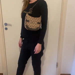 Beige Gucci axelväska med monogram - Snygg beige axelväska med klassiskt monogrammönster och mörka läderdetaljer. Väskan har en dragkedja och en dekorativ metallspänne framtill. Perfekt för att ge din outfit en lyxig touch.