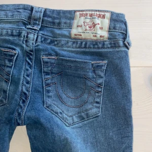 Blå jeans från True Religion - Snygga blå jeans från True Religion med klassisk femficksdesign och broderade bakfickor. De har en bootcut-stil. Dem är för små det är därför jag säljer.