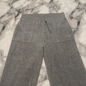 Grå mjukisbyxor från Juicy Couture - Snygga grå mjukisbyxor från Juicy Couture med elastisk midja och snörning. Byxorna har praktiska fickor fram och bak med knappar. Perfekta för en avslappnad stil.