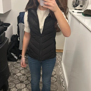 Svart Zara väst med luva  - Snygg svart dunväst med dragkedja och huva. Perfekt för kyligare dagar och ger en stilren look. Västen har en quiltad design som ger extra värme och komfort. Den är väldigt skön och  ger en en fin form. Säljer på grund av att jag har bytat stil och får inte den till användning. Original pris 600kr