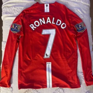 Röd Manchester United fotbollströja - Säljer en röd Manchester United fotbollströja med 'Ronaldo' och nummer 7 på ryggen. Tröjan har långa ärmar och Premier League-loggor på ärmarna. Perfekt för fans av laget och spelaren!