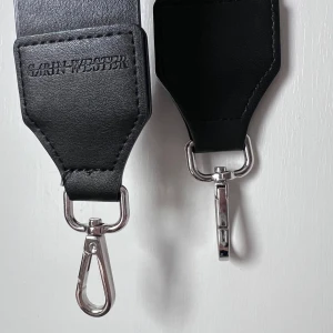 Väsk-rem från Carin Wester - Snygg svart väsk-rem från Carin Wester med silverfärgade spännen. Tillverkat i ett stilrent material som ger en modern look. Perfekt för att ge din outfit det lilla extra. Storlek: 108x5cm
