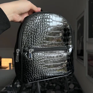 Svart ryggsäck i krokodilmönster - Snygg svart ryggsäck med krokodilmönster och glansig finish 🐊🖤 Väskan har en praktisk dragkedja framtill och justerbara axelremmar. Perfekt för att ge din outfit en cool touch. Använd knappt en handfull gånger, finns inga tecken på användning. Bredd: 20 cm Längd: 21 cm