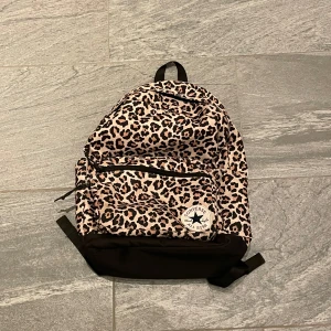 Leopardmönstrad ryggsäck från Converse - Snygg leopardmönstrad ryggsäck från Converse med svart botten och justerbara axelremmar. Perfekt för att bära dina grejer med stil. Den har en praktisk dragkedja framtill för enkel åtkomst till dina saker.
