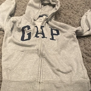 Grå hoodie från GAP Kids - Säljer en klassisk grå hoodie från GAP Kids med dragkedja och stor logga på bröstet. Tröjan har en bekväm passform med långa ärmar och en praktisk huva. Perfekt för en avslappnad stil.