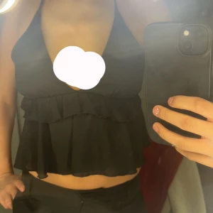 Svart volangtopp - Snygg svart topp med volanger och djup v-ringning. Toppen är ärmlös och har en croppad design som ger en trendig look. Perfekt för att styla med både byxor och kjol. Det är band man knyter vid nacken som gör att man kan spänna åt den hur man vill. Storlek Xs 