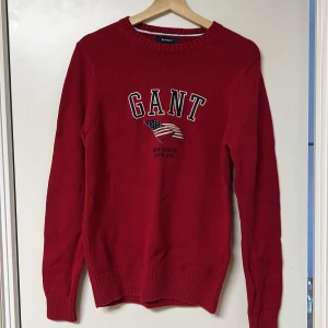 Röd tröja från GANT - Säljer en snygg röd tröja från GANT med broderad text och amerikansk flagga på bröstet. Tröjan har långa ärmar och ribbade muddar vid ärmslut och nederkant. Perfekt för en casual look!