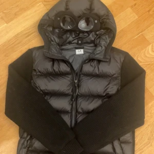 Svart Goggle Jacket från C.P. Company - Säljer en svart Goggle Jacket från C.P. Company. Jackan har en unik design med inbyggda glasögon i huvan och är tillverkad i ett glansigt material. Den har dragkedja framtill och långa ärmar i ett kontrasterande tyg.
