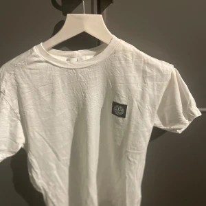 Vit t-shirt från Stone Island - Säljer en stilren vit t-shirt från Stone Island med deras ikoniska logga på bröstet. Perfekt för en avslappnad look. T-shirten är kortärmad och gjord i mjukt bomullsmaterial.