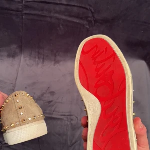 Beige mockaloafers med nitar - Snygga beige loafers i mocka från Christian Louboutin med ikoniska röda sulor. Skorna är dekorerade med guldnitar som ger en edgy look. Perfekta för att sticka ut i mängden.