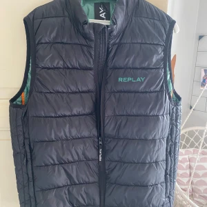 Svart dunväst från Replay - Snygg svart dunväst från Replay med dragkedja framtill. Västen har en stilren design med Replay-logga på bröstet och en kontrasterande grön insida. Perfekt för lager-på-lager under kyligare dagar.