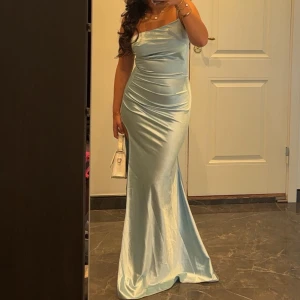 Ljusblå axelbandslös långklänning - Elegant ljusblå långklänning i satin med axelbandslös design. Perfekt för speciella tillfällen med sin smickrande passform och glansiga material. Endast använd en kväll. Skriv mer för information. 