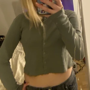 Grön långärmad kofta - Säljer en stilren grön kofta med långa ärmar och knappar framtill. Perfekt för lager-på-lager och passar till många olika outfits. Enkelt och snyggt plagg för alla tillfällen.