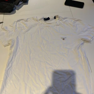 Vit t-shirt från GANT - Säljer en klassisk vit t-shirt från GANT med broderad logga på bröstet. Perfekt för en stilren och avslappnad look. T-shirten har korta ärmar och är tillverkad i mjukt material.