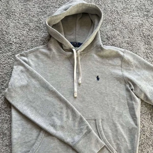 Grå hoodie från Ralph Lauren - Säljer en stilren grå hoodie från Ralph Lauren med den klassiska broderade loggan på bröstet. Tröjan har en justerbar huva med snören och en bekväm passform. Perfekt för en avslappnad look. Den är i toppskick!