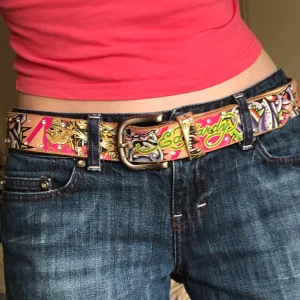 Rosa Ed Hardy bälte - Ca 110 cm! Jättesnyggt rosa ed hardy bälte. Coola detaljer, gems/rhinestones längs med hela bältet. Zooma för detaljer! Bältet har tecken på användning med det är kosmetiskt och det är inte trasigt eller så. Man kan se på bilden om man zoomar in där det rosa trycket är slitet.