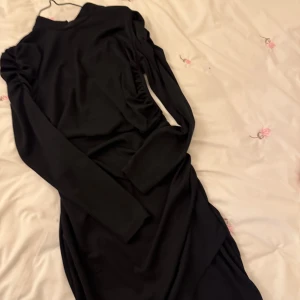 Svart långärmad klänning - Snygg svart klänning med långa ärmar och en elegant drapering på sidan. Perfekt för en stilren look. Klänningen har en figurnära passform och är tillverkad i ett mjukt material som ger en bekväm känsla. Använd bara en gång. 