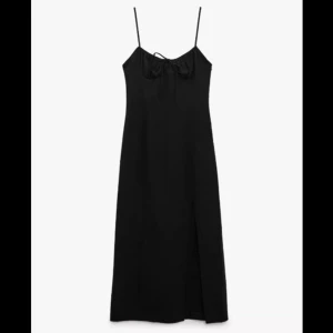 Svart långklänning med smala axelband - Elegant svart långklänning med smala axelband och en subtil slits framtill. Klänningen har en knytning vid bysten som ger en fin detalj. Perfekt för en stilren look.