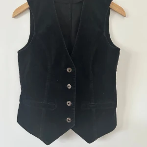 Svart manchester väst från Kappahl - Snygg svart manchester väst från Kappahl med knappar framtill. Perfekt för att ge din outfit en stilfull touch. Västen har en klassisk design med två dekorativa fickor.