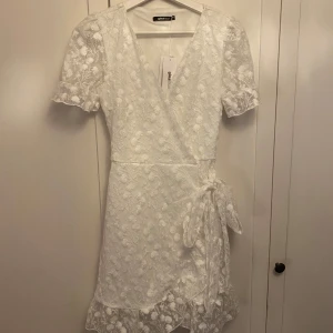 Vit studentklänning med spets från Gina Tricot - Säljer en superfin vit studentklänning med spets från Gina Tricot i omlottmodell. Klänningen har korta ärmar och ett fint blommigt mönster. Den knyts i midjan och har en volangdetalj vid fållen. Perfekt för sommarens alla tillfällen! 🌸Endast provad en gång med etiketten kvar, alltså i helt nytt skick!!! Original pris 700kr
