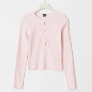 Ljusrosa tröja med rosetter - Säljer en söt ljusrosa ribbad tröja från Gina. Tröjan har långa ärmar och en unik knytning framtill som ger en fin detalj. Perfekt för en stilren look.