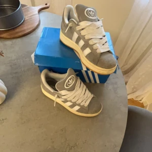 Grå och vita Adidas campus - Säljer ett par snygga grå och vita sneakers från Adidas med klassiska ränder och bred snörning. Skorna har en gummisula i brunt och kommer i originalkartong. Perfekta för en avslappnad stil.