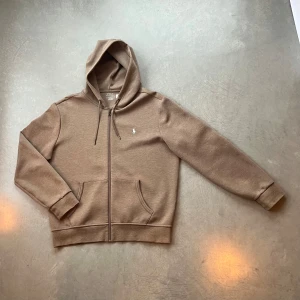 Ralph Lauren Beige zip hoodie - Säljer en snygg beige hoodie från Ralph Lauren med dragkedja och klassisk logga på bröstet. Perfekt för en avslappnad stil. Den är använd några få gånger och är i väldigt bra skick. Inköpt för ca 2200kr för ungefär 1,5 år sedan. Passar bra. 