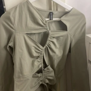 Grön topp från Divided - Trendig grön topp från Divided med unika snörningsdetaljer och cut-outs framtill. Toppen har långa ärmar och en stilren design som passar perfekt för en modern look.  HAR ALDRIG ANDVÄNT