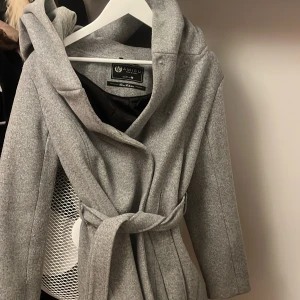 Grå kappa med knytband - Elegant grå kappa från Alaïa med knytband i midjan och en stor huva. Perfekt för kyligare dagar med sin stilrena design och mjuka material.